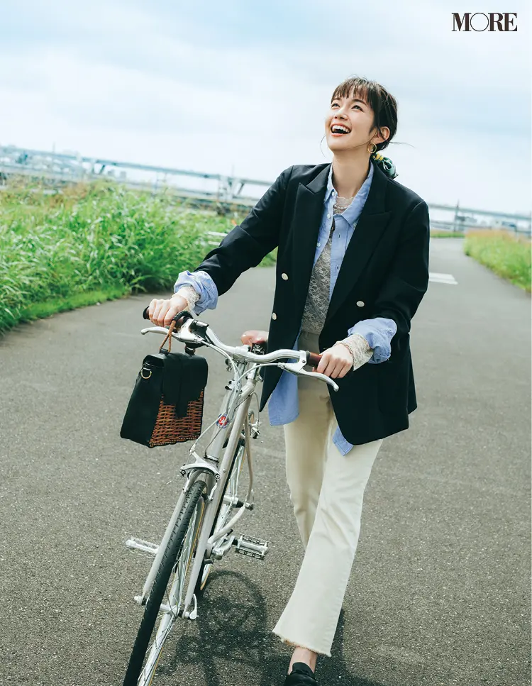 レーストップスとシャツにネイビージャケットをはおり、ホワイトデニムをはいて自転車を押す佐藤栞里