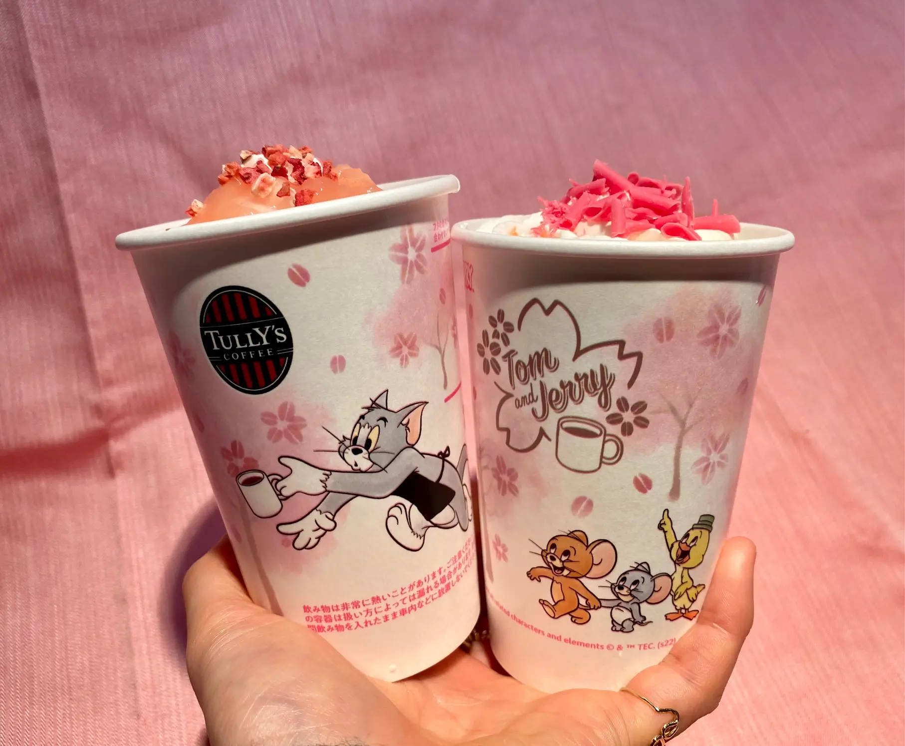【タリーズ 新作】「トムとジェリー 桜舞うスペシャルコーヒータイム」ドリンク「トムとジェリー 桜舞う苺カフェラテ」、「トムとジェリー &TEA 桜香る桃のティーオーレ」を手に持っている様子